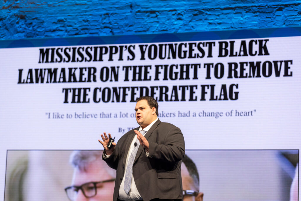 Speaker presenting on stage in front of a projected slide with a large headline.