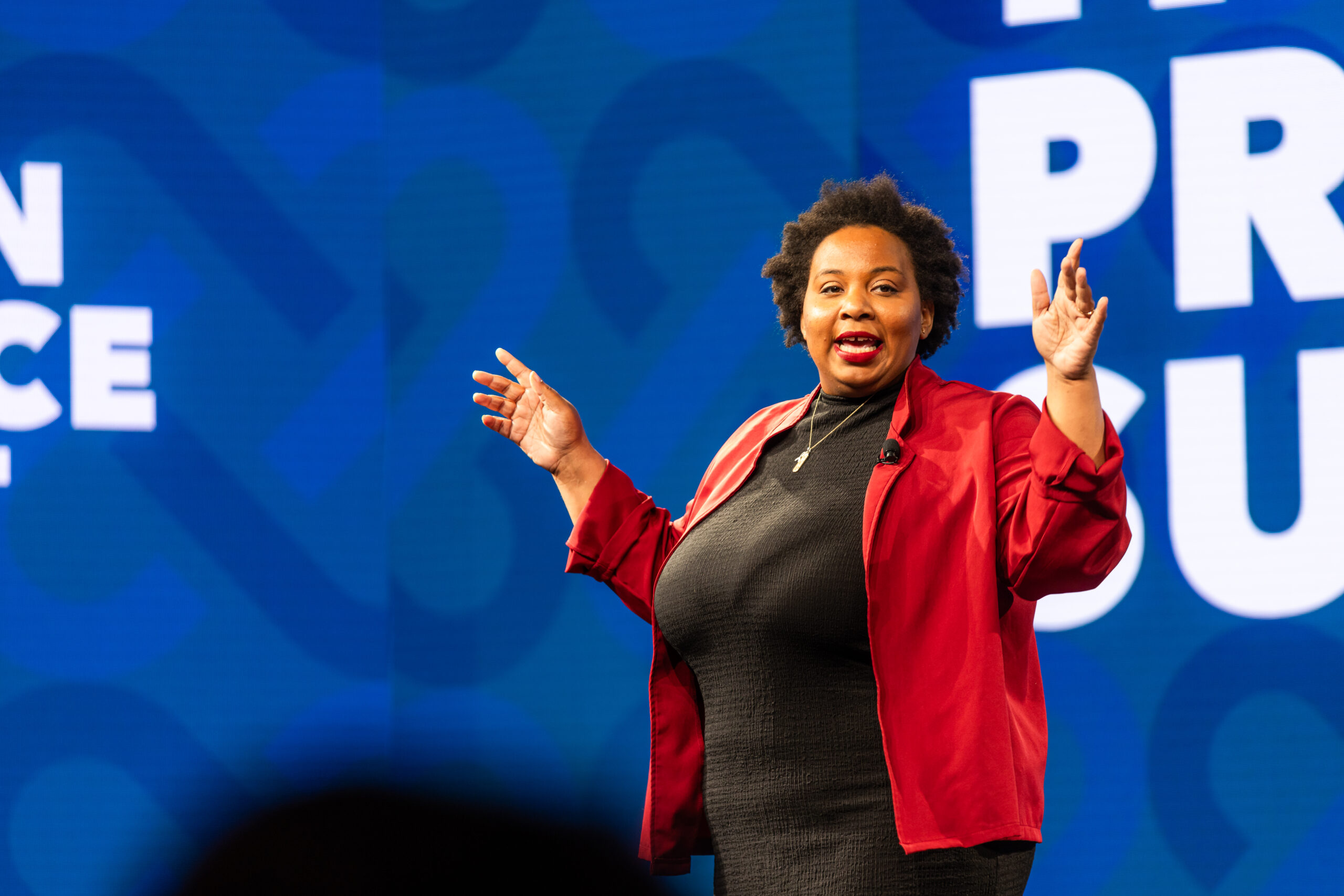 Speaker in a red jacket gesturing on stage in front of a blue backdrop.
