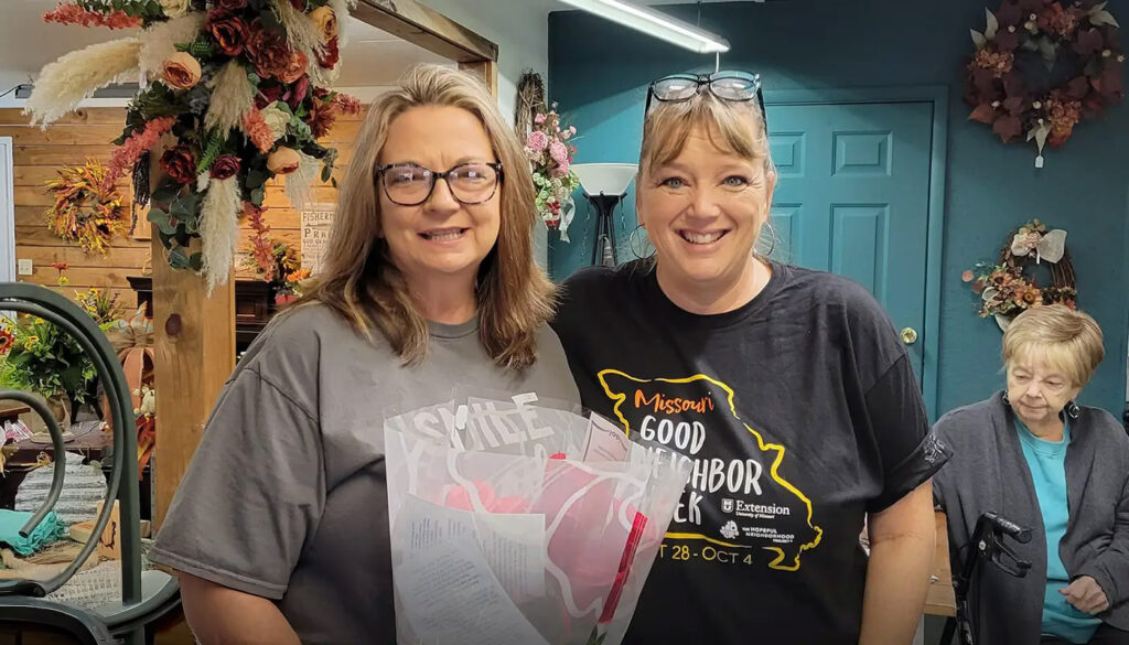 Two women smiling indoors, holding a bouquet and gift bag.