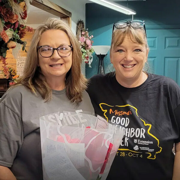 Two women smiling indoors, holding a bouquet and gift bag.