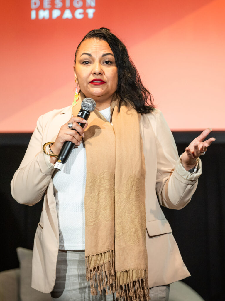 Woman holding a microphone speaking on stage against a red backdrop.