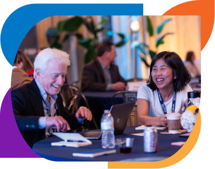 Two conference attendees sitting at a table smiling and talking, with notebooks, drinks, and laptops in front of them.